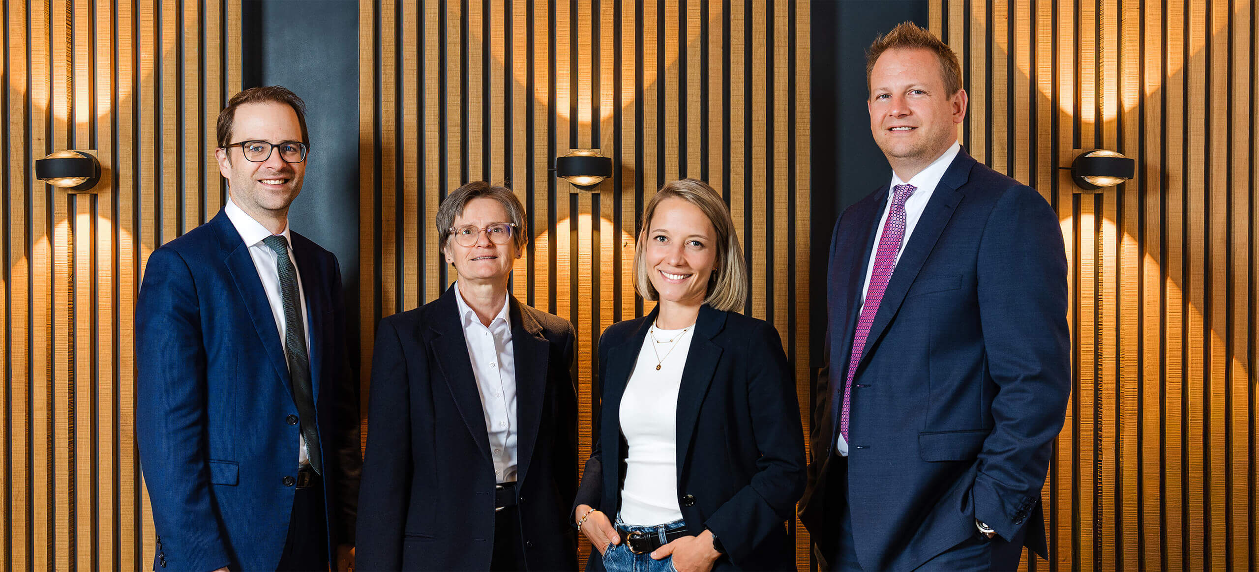 Die Partner von Glensk + Partner stehen zusammen vor einer Holzwand.