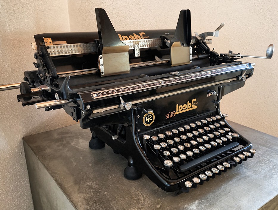 Auf dem Bild ist eine große, schwarze mechanische Schreibmaschine zu sehen. Sie steht auf einer glatten, grauen Oberfläche vor einer hellen Wand. Das Modell trägt die Aufschrift „Ideal Blitz“ in goldfarbener Schrift. Vorne befindet sich eine klassische Tastatur mit runden Tasten in Schwarz und Weiß. Oberhalb der Tastatur sind zwei Papierhalter und mehrere metallene Hebel und Walzen erkennbar, die zum Einspannen und Transportieren des Papiers dienen.