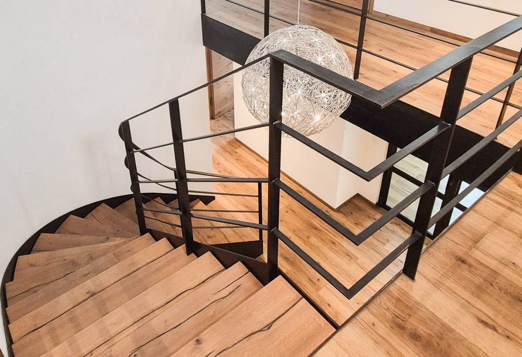 Ein modernes Treppenhaus in einem Innenbereich mit Holzstufen und einem schwarzen Metallgeländer. Die Treppe ist gewendelt und führt von einem oberen Stockwerk nach unten. Der Boden besteht ebenfalls aus Holz mit natürlicher Maserung. Über dem Treppenauge hängt eine große, kugelförmige Lampe aus feinen, weißen Drähten, die an ein Kunstobjekt erinnert. Die Wände sind weiß und schlicht gehalten, wodurch das Design der Treppe besonders hervorsticht.